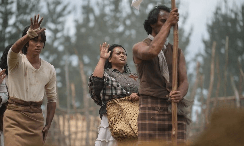 Three people in historical attire stand outdoors, raising their right hands. The woman in the center carries a woven basket. The background shows trees and wooden poles, suggesting a rural or historical setting.