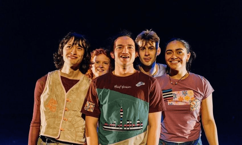 Five young adults stand close together, smiling at the camera. They are brightly lit against a dark background and wear colorful, casual clothing.