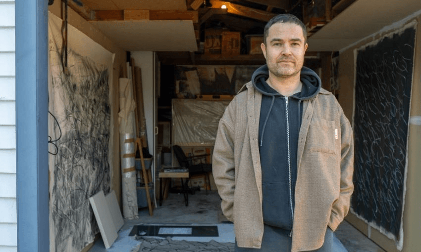 A man stands in front of a garage art studio, wearing a brown jacket and black hoodie. Behind him are large abstract artworks, canvases, and art supplies.