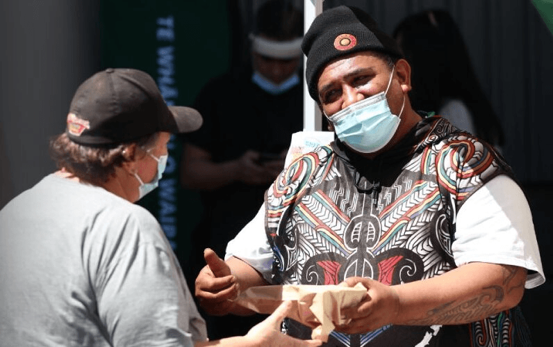Two people wearing masks talk outside. One wears a hat and patterned vest, handing food in a paper tray to the other. “THE BULLETIN” is written vertically on the left side.