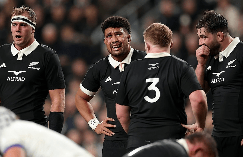 four men in black rugby uniforms stand, puffed, with a large crowd behind them