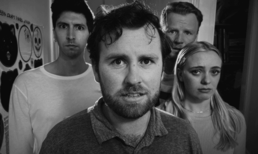 Four adults stand together indoors, facing the camera with serious or concerned expressions. The image is in black and white, and the background includes a wall with posters and a shelf.