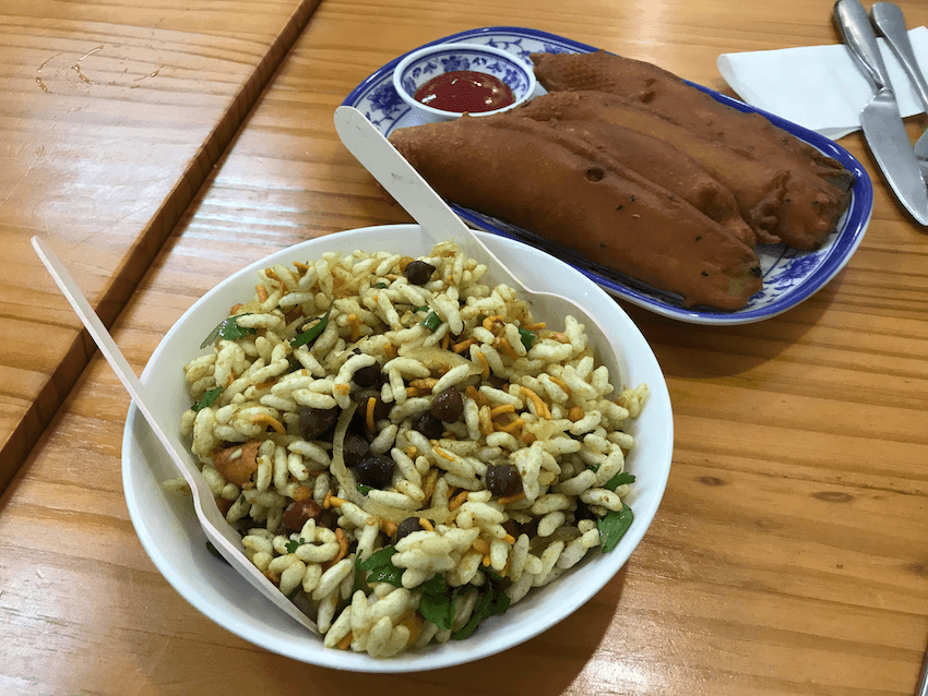 A bowl of puffed rice snack with vegetables sits next to a plate of fried snacks and ketchup on a wooden table, with utensils in the background.
