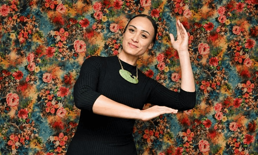 A woman in a black dress poses with her arms raised against a bright floral-patterned background. She wears a large green pendant necklace and smiles slightly at the camera.