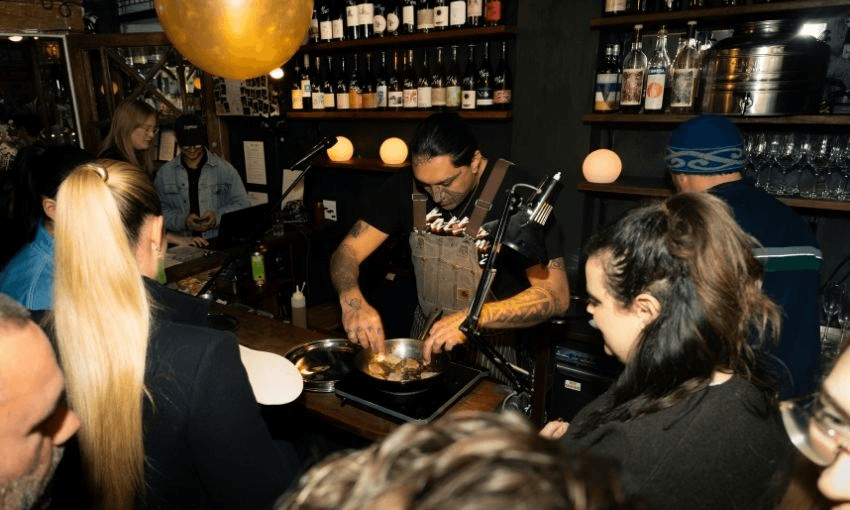 A chef in overalls prepares food at a counter while several people watch in a cozy, dimly lit bar or restaurant with bottles on shelves behind him.
