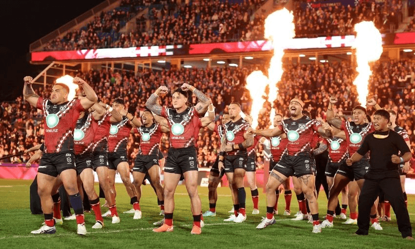 A rugby team in red and black performs a pre-match haka dance on a field, with flames shooting up behind them and a large crowd watching from the stands.
