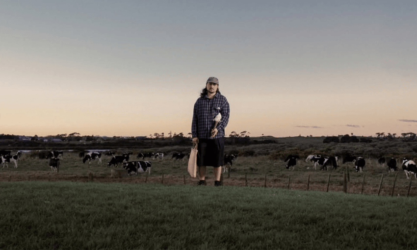 A person in casual clothes stands on grassy land holding a paddle, cows graze in the background under a dusk sky.