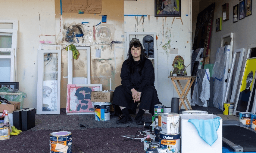 A person in black clothes sits in an art studio surrounded by paint cans, artworks, and art supplies, with colorful paintings and splattered walls in the background.