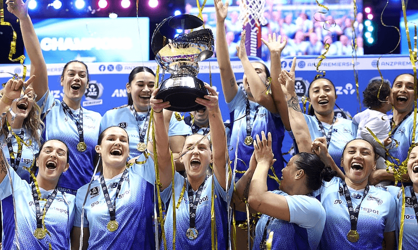 a close up group shot of a netball team of women in blue uniforms celebrating wearing medals and one holding a large trophy as streamers fall