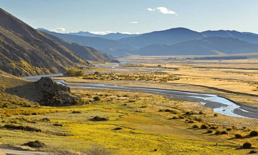 Sunny valley landscape with a winding river, grassy fields, and yellow patches. Hills rise on the left, mountains line the horizon, and the sky is mostly clear with a few clouds.