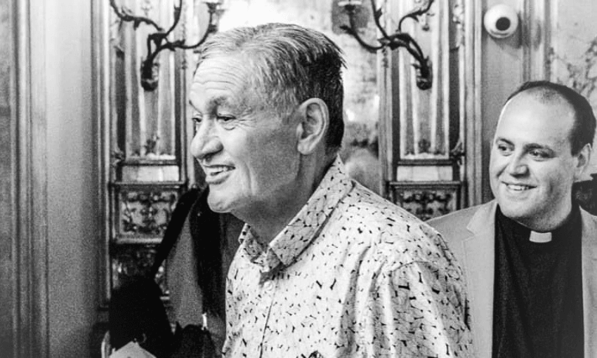 Two men smile and walk together in an ornately decorated room. The man in front wears a patterned shirt; the man behind him wears a clerical collar. The atmosphere appears joyful and warm.
