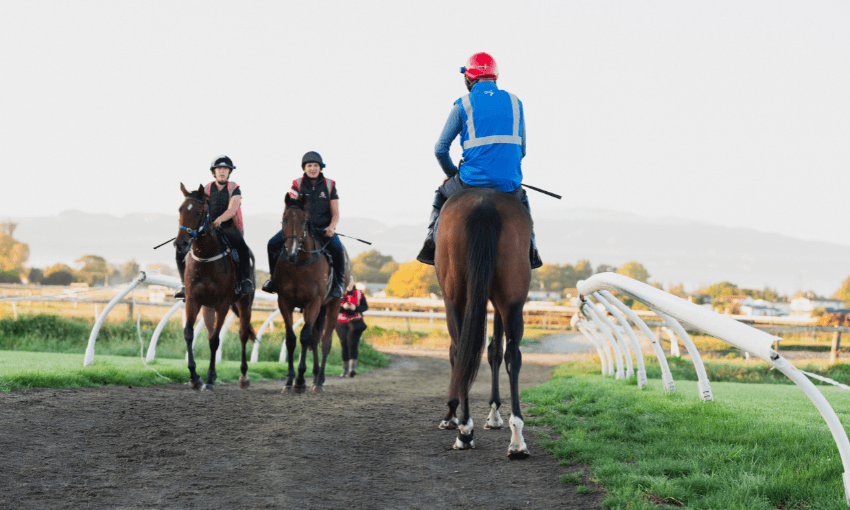 A photo of horses and jockeys on a race track. The sky is dull and cloudy.
