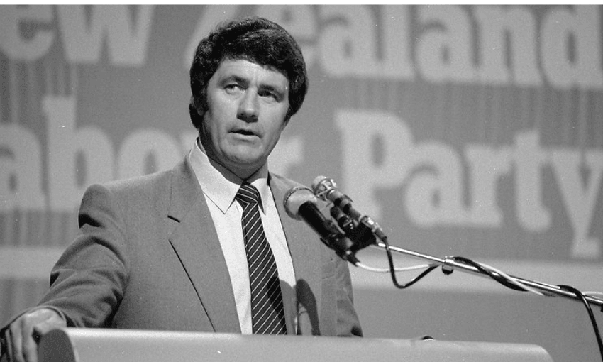 A man in a suit and tie speaks at a podium with microphones. A blurred sign behind him reads “New Zealand Labour Party.” The scene appears to be at a formal event or conference.