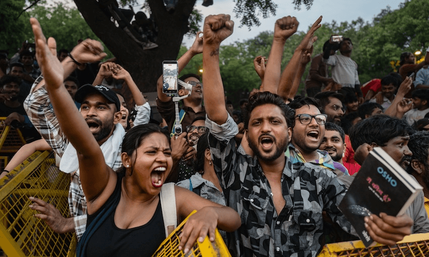 A group of people stand behind a yellow barricade, raising their fists and shouting passionately. One person holds a phone, another holds a book. Trees are visible in the background.
