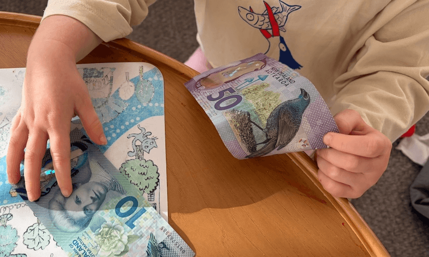 A child holds a New Zealand $50 note in one hand and touches a $10 note on a wooden table, next to a coloring sheet.