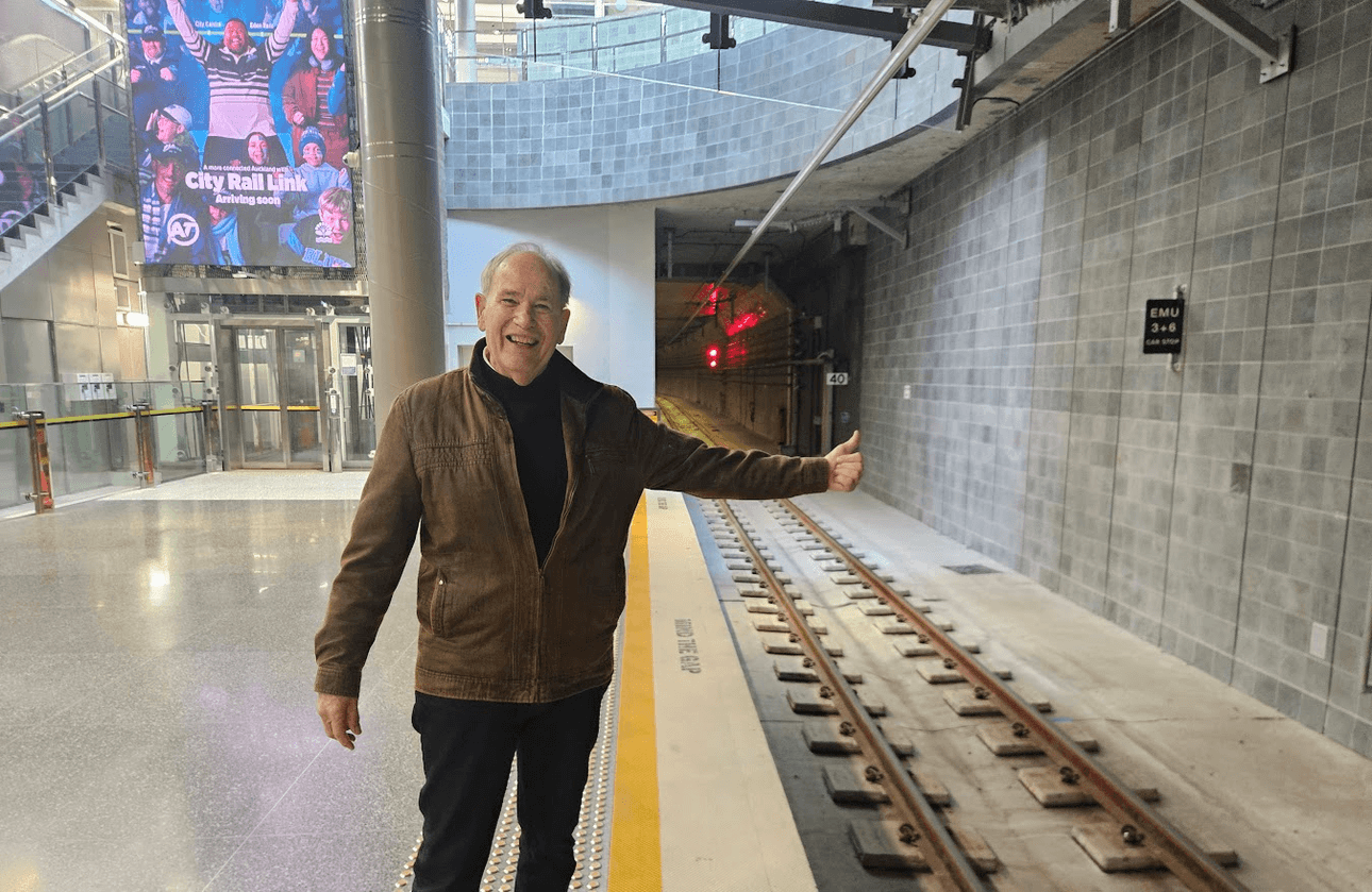 A smiling man in a brown jacket stands near train tracks in an indoor station, giving a thumbs-up. Behind him is an entrance and a large poster with people and the text “City Rail Link.”.