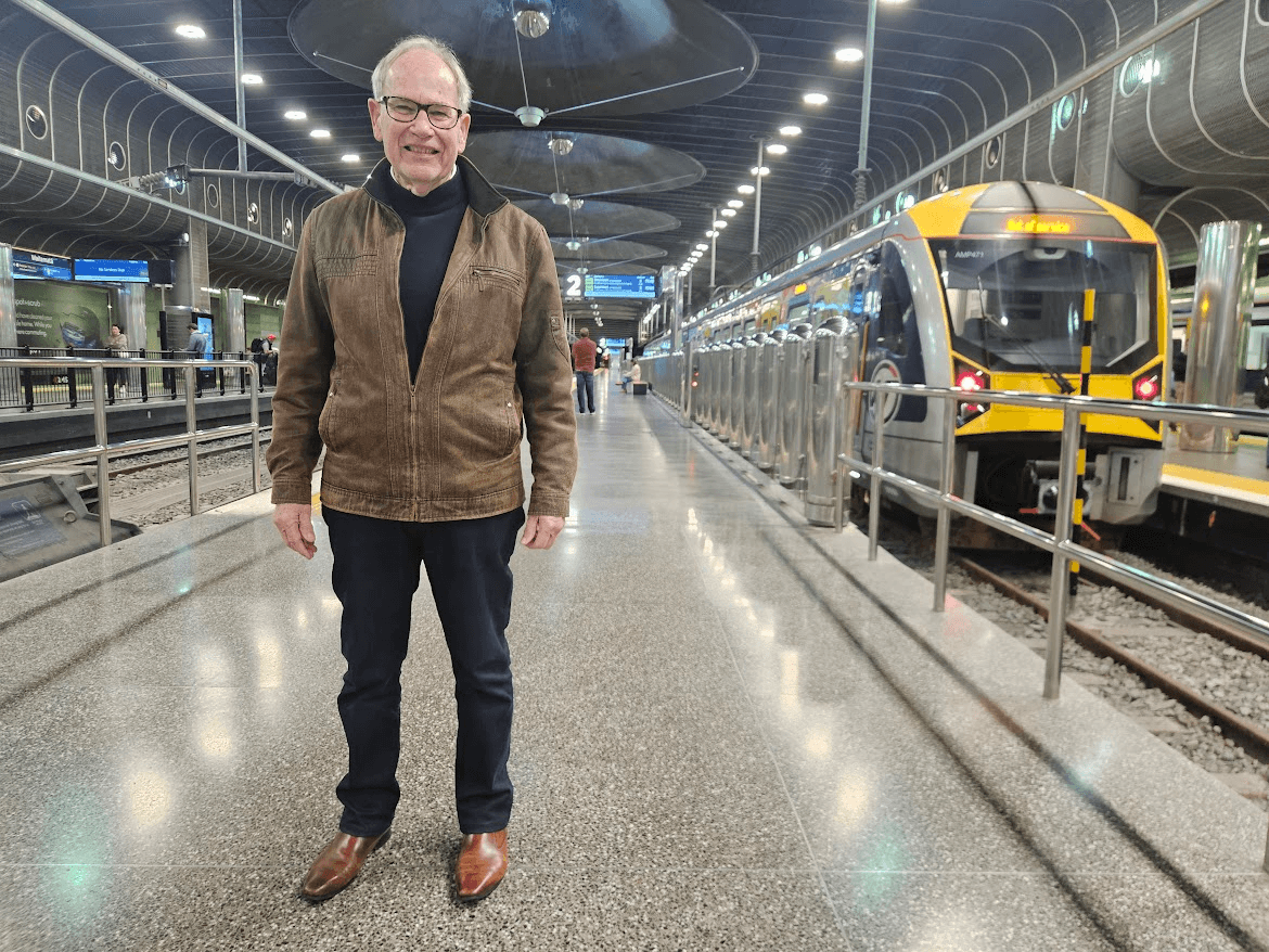 An older man wearing glasses and a brown jacket stands smiling on a shiny train platform next to a modern yellow-and-grey train inside a well-lit underground station.