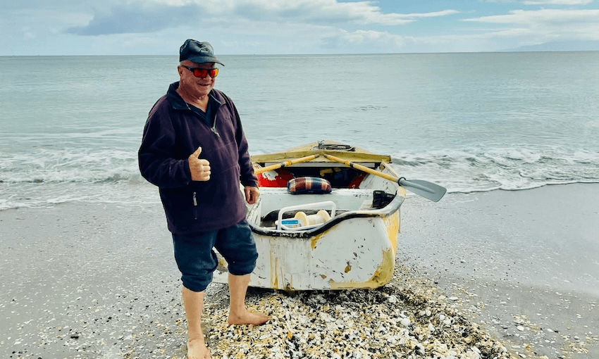 A barefoot man in a cap and sunglasses stands on a shell-covered beach, smiling and giving a thumbs-up beside a small rowboat with oars and supplies, with the ocean and cloudy sky in the background.