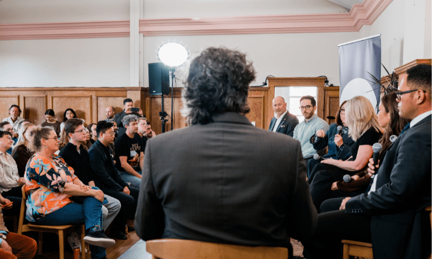 A group of panelists speaks to an audience in a wood-paneled room. People are seated, listening attentively. A man with curly hair is in the foreground, seen from behind. Bright lights and a banner are in the background.