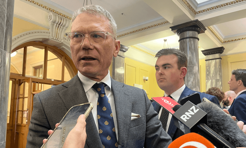 A man in a suit speaks to reporters holding microphones and phones in a formal building with columns and ornate ceilings. Another man stands behind him.