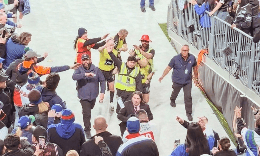 A crowd surrounds security escorting a man off a sports field. Stewards in yellow vests and staff in dark uniforms guide the man as fans watch and take photos from the stands.