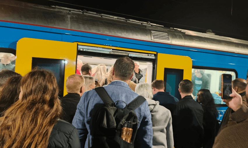 A crowd of people waits to board a blue and yellow train at night. The train doors are open, and passengers are visible inside. Some people are looking at their phones.