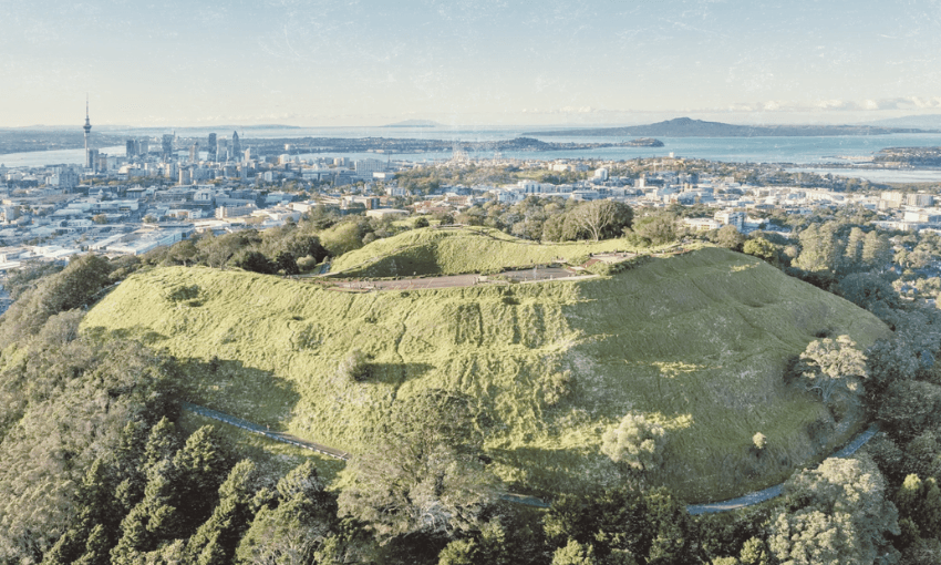 An aerial shot of Maungawhau / Mt Eden looking towards downtown Auckland. The cityscape is in the background, including the sky tower, Waitematā Harbour and Rangitoto.