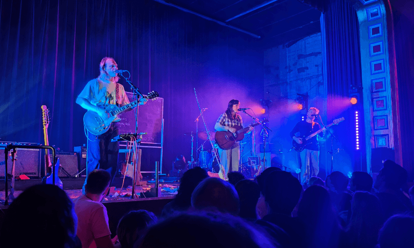 A band performs on a dimly lit stage with blue and purple lights, playing guitars and singing. The audience watches from below, and musical equipment is visible around the musicians.