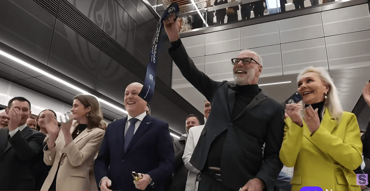 A group of people in formal attire celebrate indoors; a smiling man in glasses and black holds up a cut ribbon while others around him applaud enthusiastically.