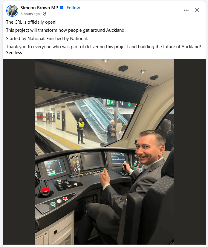 A man in a suit sits in the driver’s seat of a modern train cab, smiling and giving a thumbs up, with train controls and station visible through the front window.