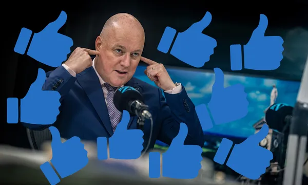 A man in a suit covers his ears with his fingers while surrounded by multiple blue thumbs-up icons, sitting at a desk with microphones and a blurred background.