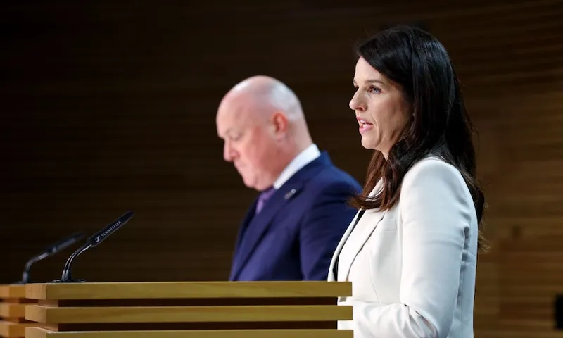 A man in a blue suit stands in the background as a woman with long dark hair and wearing a white blazer speaks at a lectern