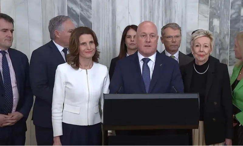 A group of professionally dressed people stand together indoors, with one man speaking at a podium, flanked by others. Marble-patterned walls are visible in the background.