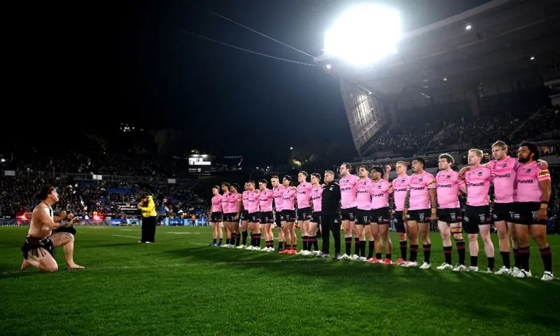 A rugby team in pink jerseys stands in a line on the field at night, while a person kneels in front performing a traditional ceremony. Stadium seats and spectators are visible in the background.