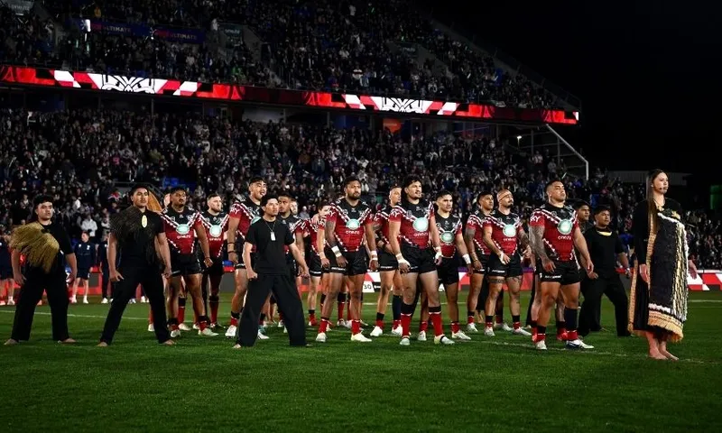 A rugby team in black and red uniforms performs a haka on a field, joined by others in traditional attire, with a stadium full of spectators in the background.