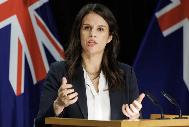 A woman in a dark blazer and white shirt speaks at a podium with microphones. New Zealand flags are visible in the background. She gestures with her hands while addressing an audience.