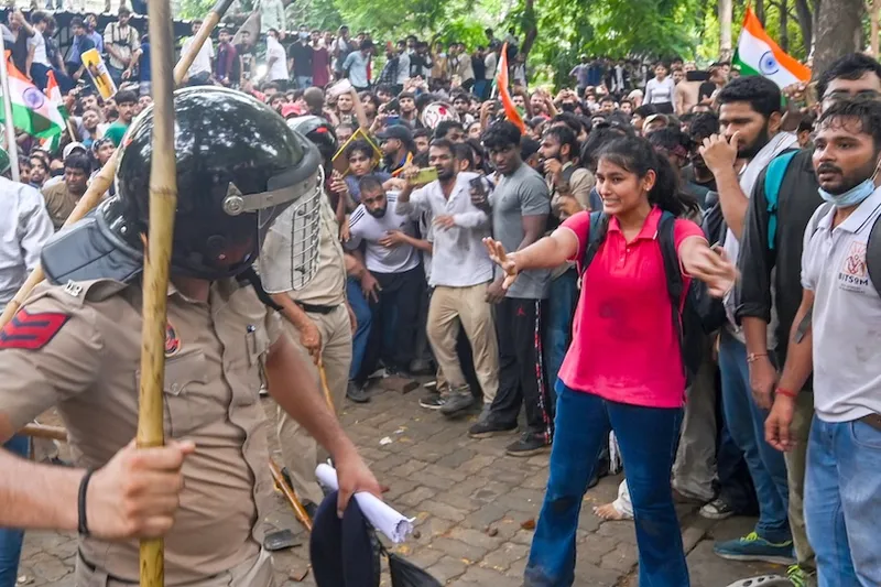 A large crowd protests outdoors; a police officer with a baton faces a woman in a pink shirt holding up her hands. Some people wave Indian flags in the background. Trees and paved ground are visible.