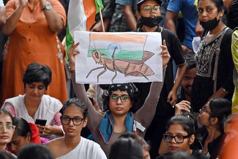 A person holds a drawing of an insect over an Indian flag at a crowded protest, surrounded by people, some wearing glasses and masks, with serious expressions.