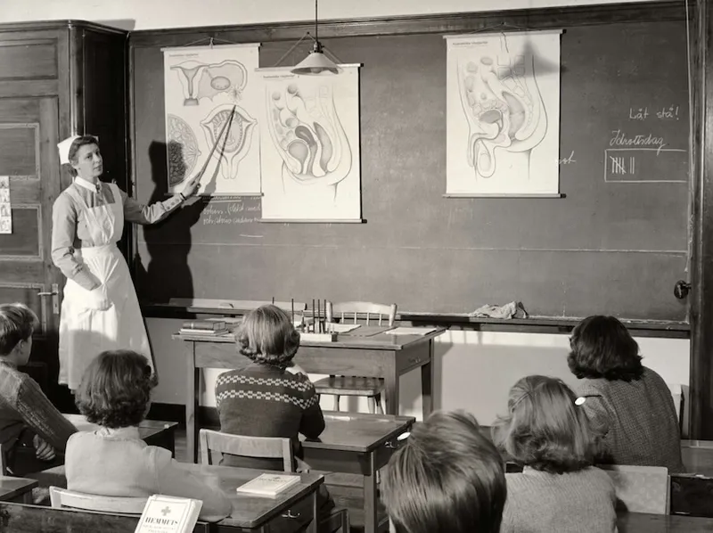 A woman in a dress and apron points to anatomical diagrams on a chalkboard while a classroom of young students watch and listen attentively.