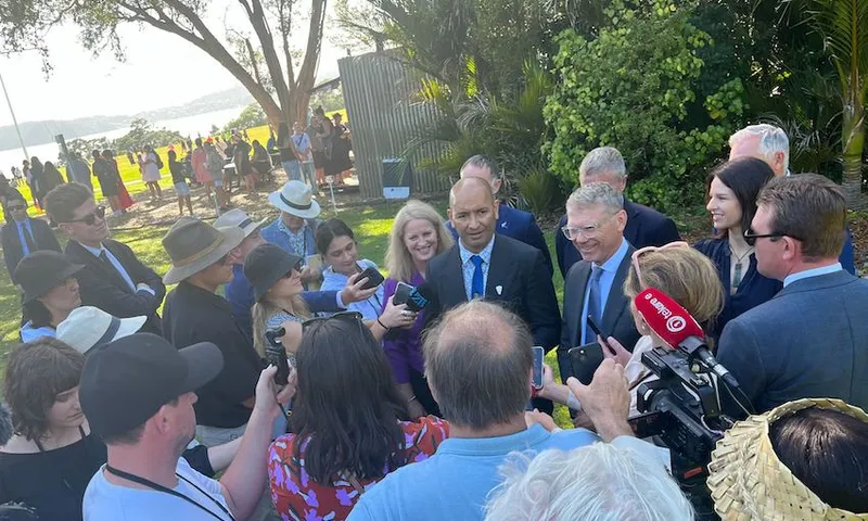 A group of people, including media personnel with cameras and microphones, are gathered around two men wearing suits in an outdoor setting. The background features trees and a body of water with more people in the distance.