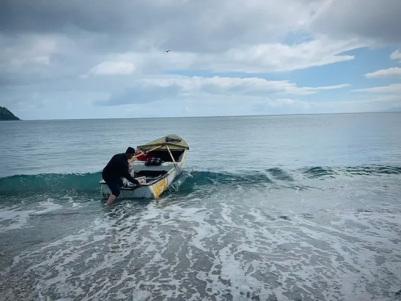 A person pushes a small, weathered boat into the gentle waves of a calm sea under a cloudy sky, with distant land visible on the left.