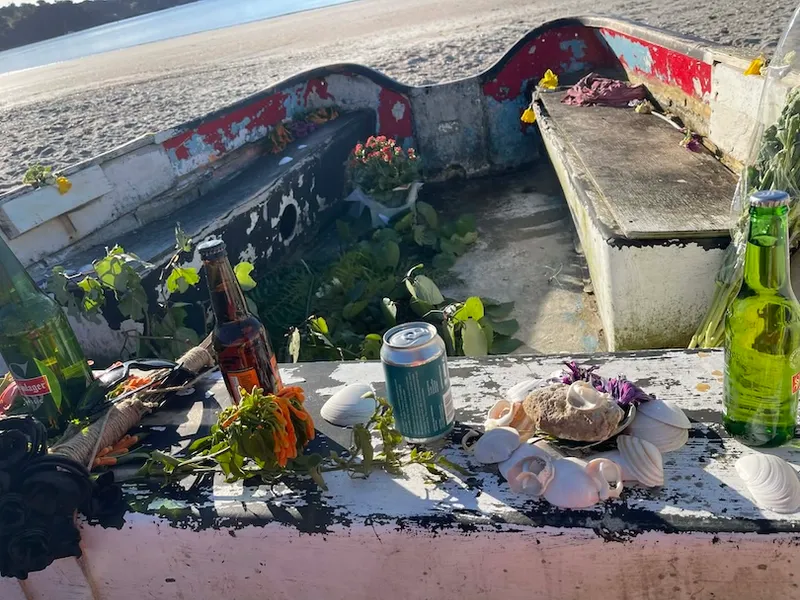 A weathered boat is filled with flowers, empty bottles, shells, and a can, resting on a sandy beach with the sea in the background. Sunlight casts shadows across the scene.