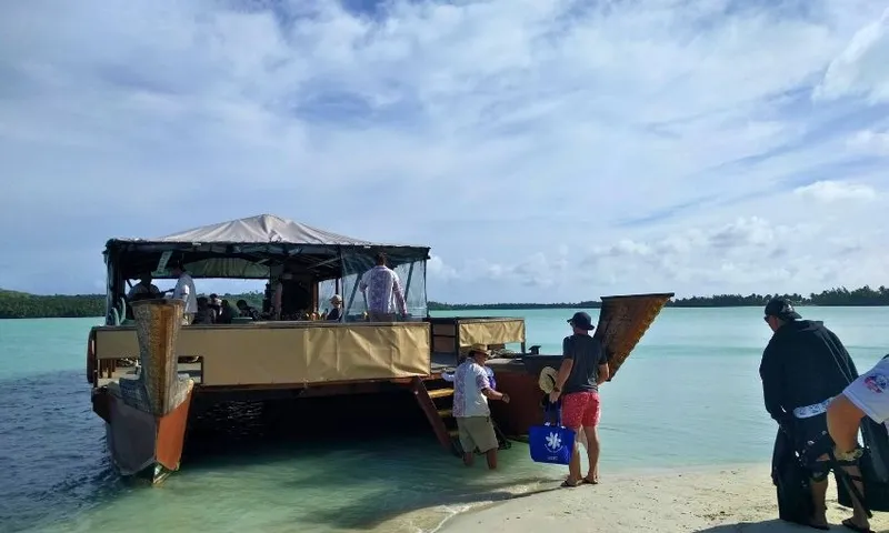 Several people board a large wooden boat from a sandy beach, with clear turquoise water and a partly cloudy sky in the background.