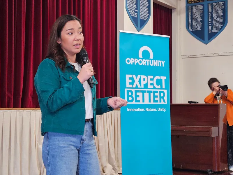 A woman speaks into a microphone in front of a blue banner that reads “EXPECT BETTER.” A person in orange stands near a piano taking a photo. Red curtains and wall plaques are visible in the background.
