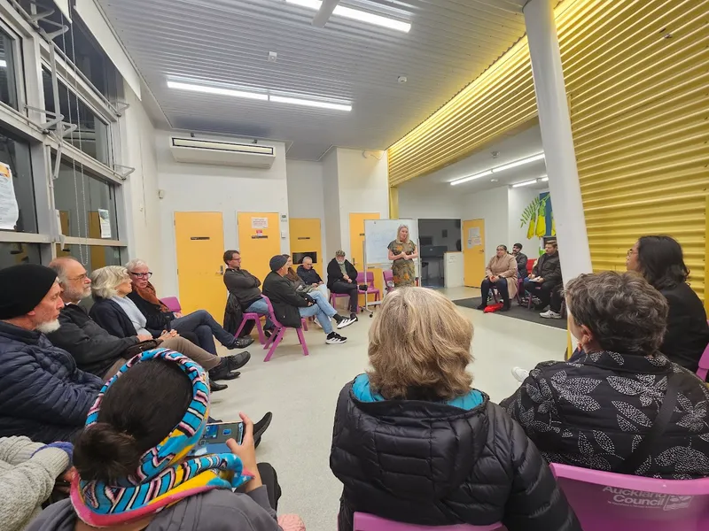A group of people seated in a circle indoors listen to a woman speaking at the front. The room has yellow and white walls, and some participants wear winter clothes.