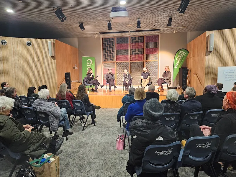 A panel of five people speaks onstage to an audience in a modern room with wood-paneled walls and patterned art behind the speakers. The seated audience listens attentively.