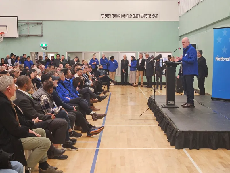 A man in a blue jacket speaks at a podium to a seated audience in a gymnasium. People stand along the wall, and a sign above reads “FOR SAFETY REASONS: DO NOT KICK OBJECTS AGAINST THIS WALL.”.
