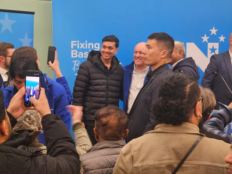 A group of people are gathered indoors, some posing for a photo, while others take pictures with their phones. Blue banners with text and stars are visible in the background.