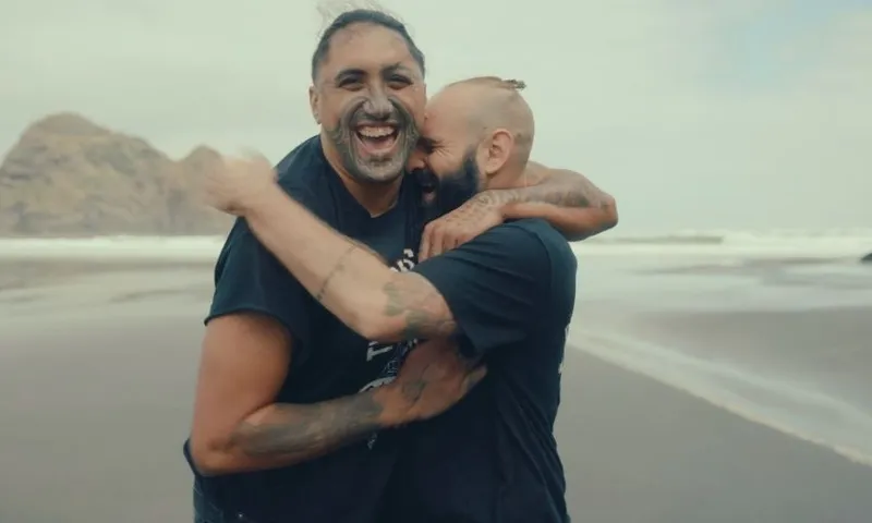 Two people with tattoos and black shirts laugh and embrace on a sandy beach with rocky cliffs and ocean waves in the background under a cloudy sky.
