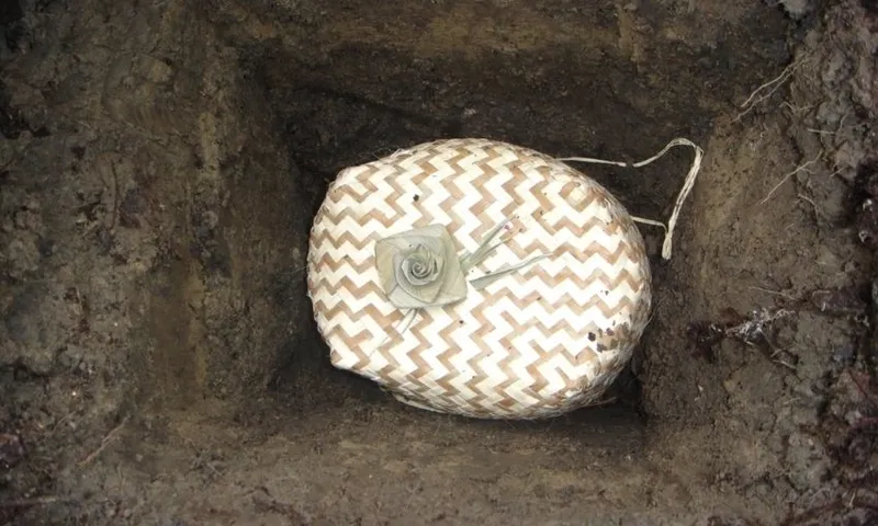 A woven, oval-shaped container with a white rose on top lies in a dirt grave, viewed from above.